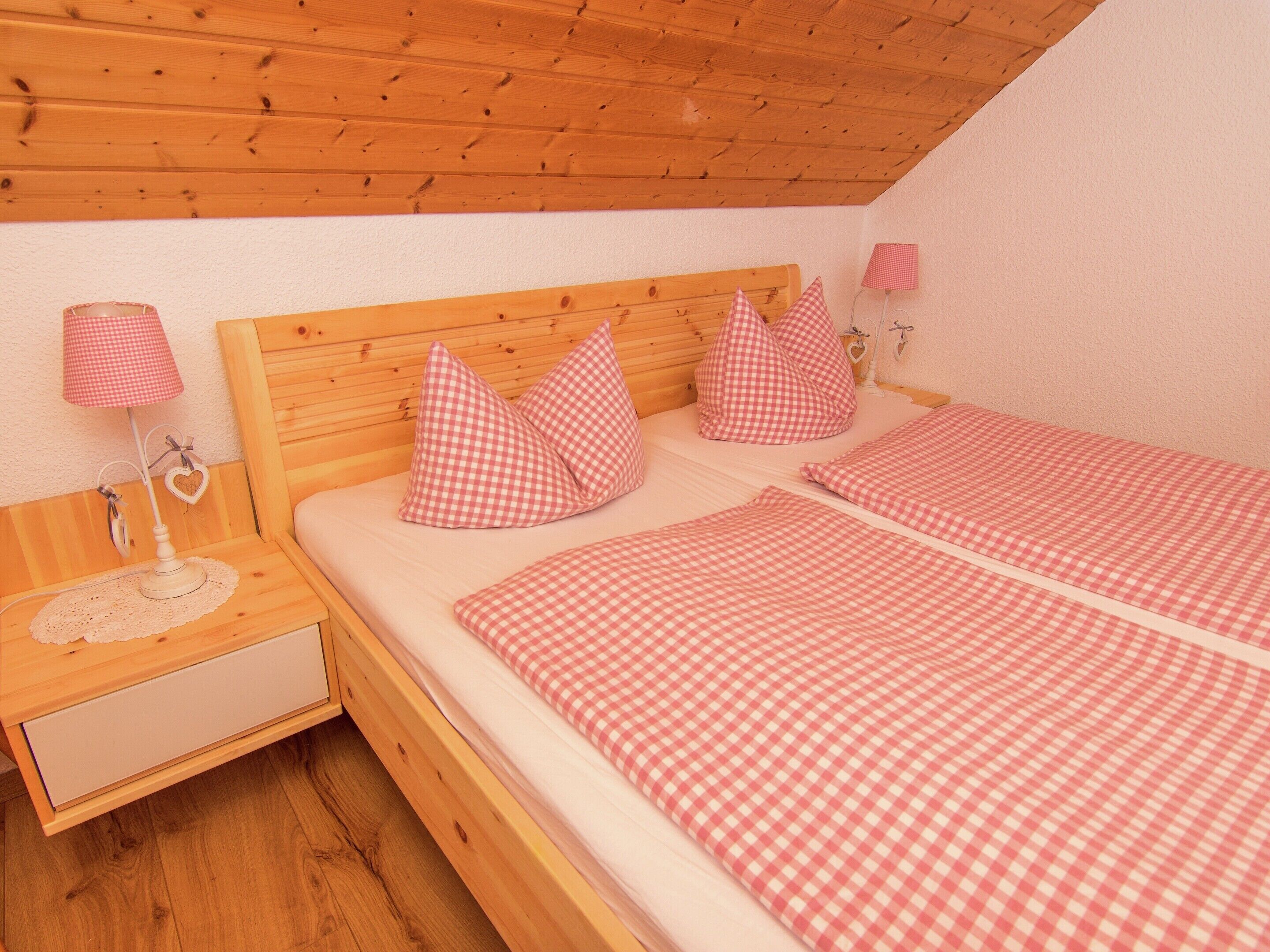 Un dormitorio con cabecera de madera, dos camas con ropa de cama a cuadros, una mesita de noche con lámpara y una mesita auxiliar con adorno con forma de corazón.