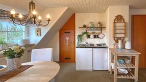 A compact kitchen with white cabinets, a sink, and a stove. There is a chandelier above the dining table, which has a white tablecloth. A small plant is on the windowsill, and a wooden shelf holds kitchen items. - Holiday Home With Garden in Weissenbrunn (Weissenbrunn)
