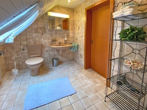 A bathroom with a toilet, sink, and shower area. There is a blue mat on the floor and a metal shelf with towels and a hairdryer.