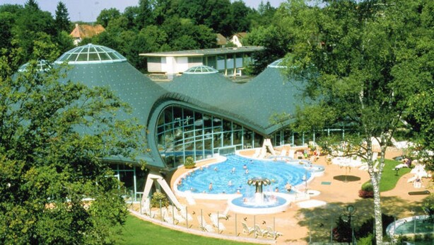 A swimming pool area with a large water slide and a fountain, surrounded by trees and a building with a unique curved roof. - Apartment in Bad Durrheim Near Lake Constance (Bad Duerrheim)