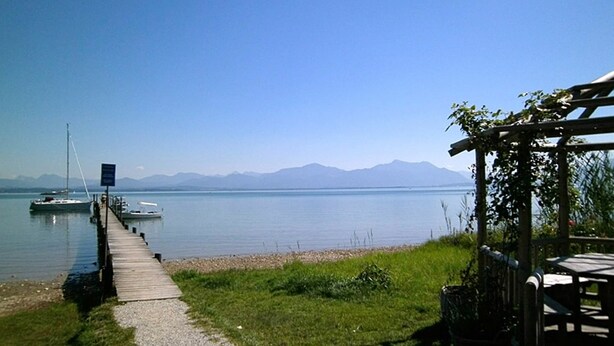 Vista del lago con un muelle de madera que lleva a los botes, una zona de picnic y montañas al fondo.