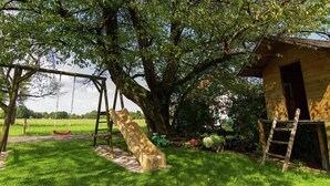 Un parque infantil con un tobogán y columpios, un árbol y una pequeña estructura de madera.