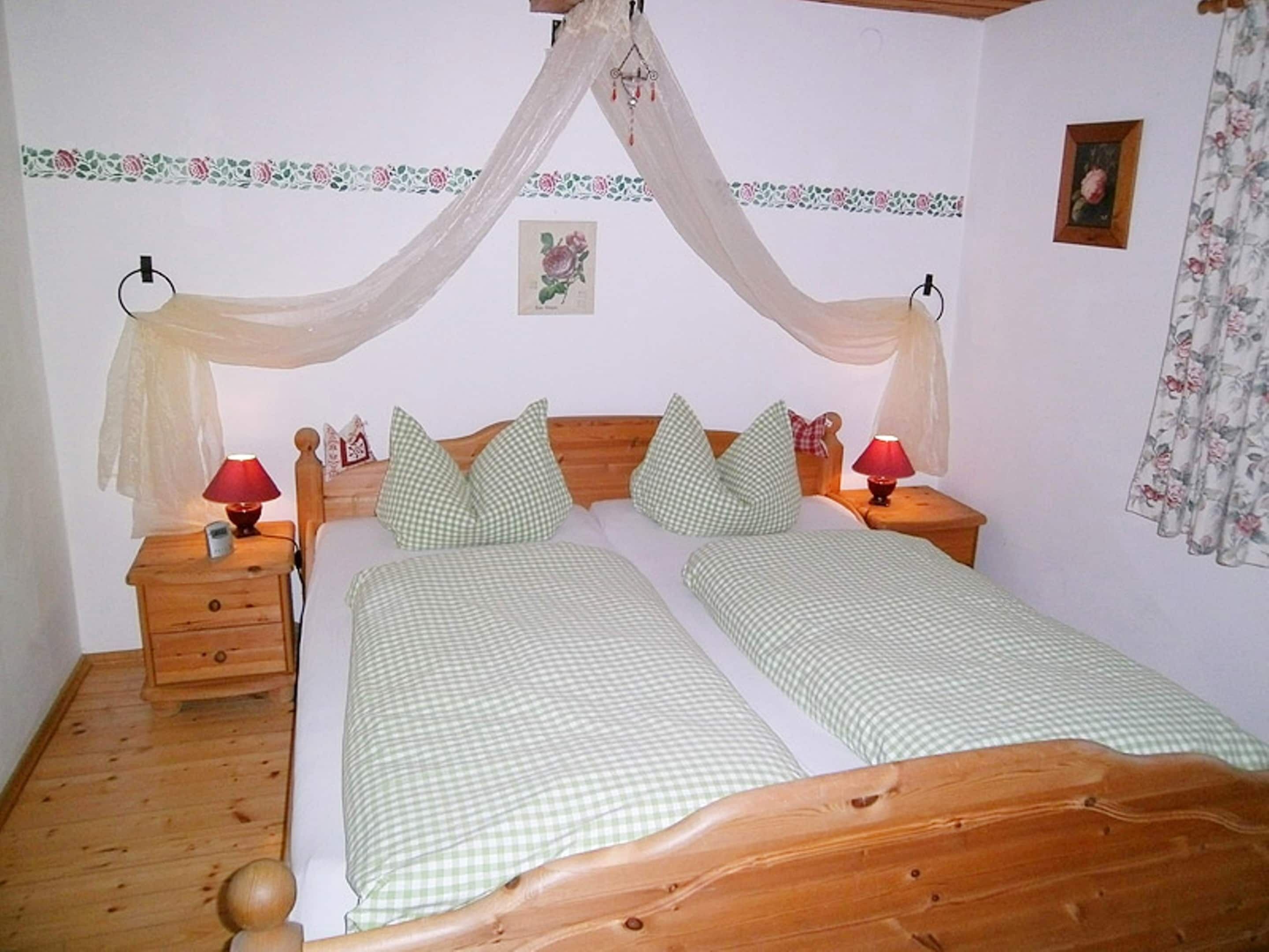 Un lit double avec une tête de lit en bois, deux tables de chevet avec des lampes, un rideau à motifs floraux et un petit tableau encadré accroché au mur.