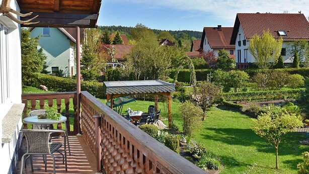 Un balcón con mobiliario exterior, vista a un jardín verde y casas al fondo.