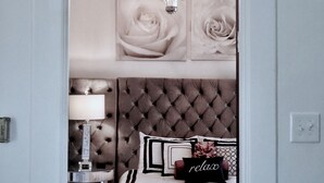 Un dormitorio con cabecera capitonada, una cama con cubrecama blanco, una mesita de noche con lámpara, y una pieza decorativa sobre la cama.