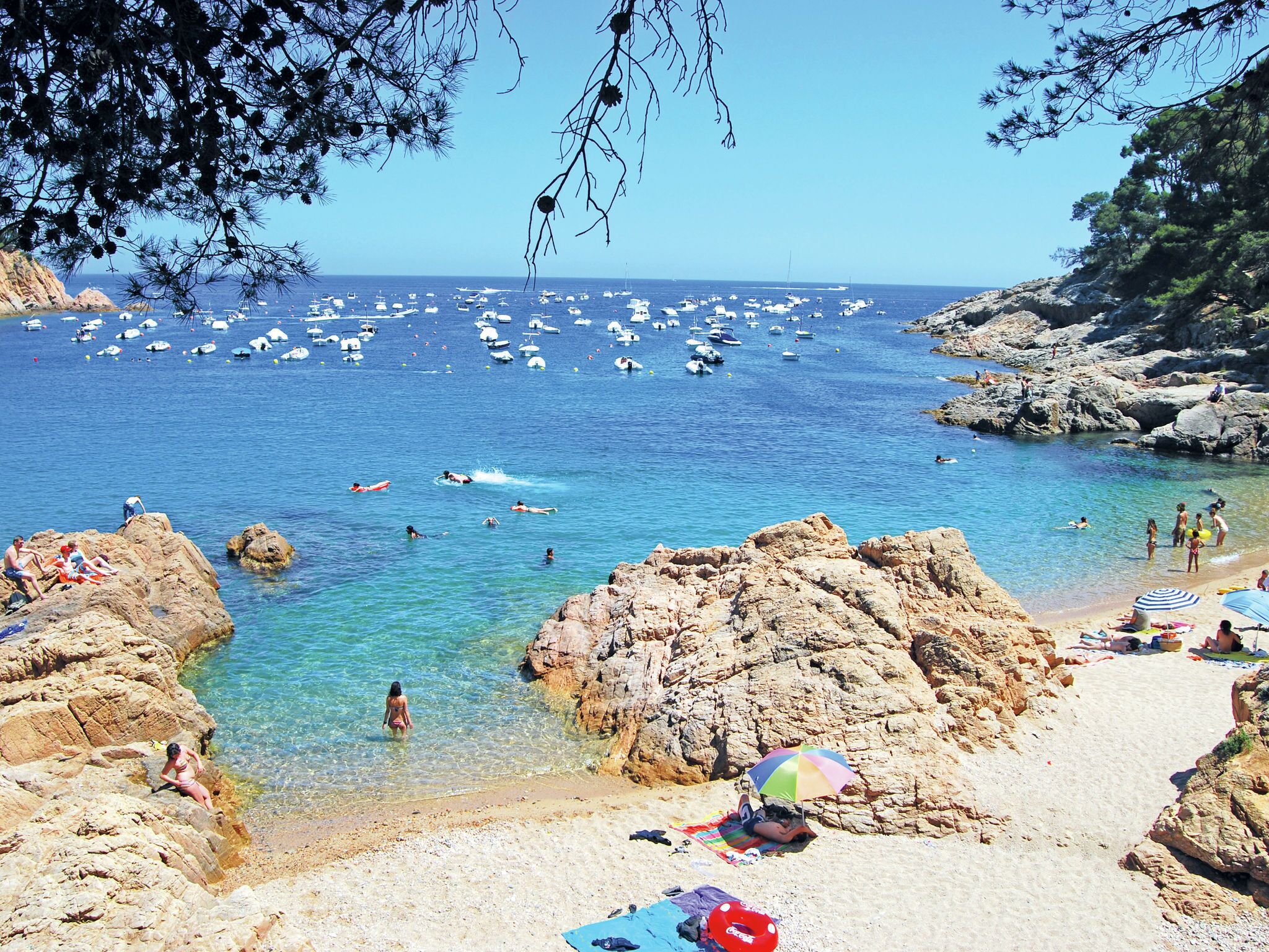 Una playa costera con terreno rocoso, agua azul cristalina y numerosos barcos.