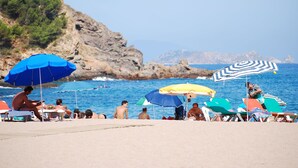 Una playa con gente tomando el sol y nadando, rodeada de acantilados rocosos y el mar.