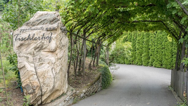 A stone sign with 'Appartments Tischlerhof' inscribed, a paved pathway, and a canopy of trees. - Holiday Apartment "Tischlerhof - Typ B" with Wi-Fi, Terrace and Mountain View (Dorf Tirol bei Meran)