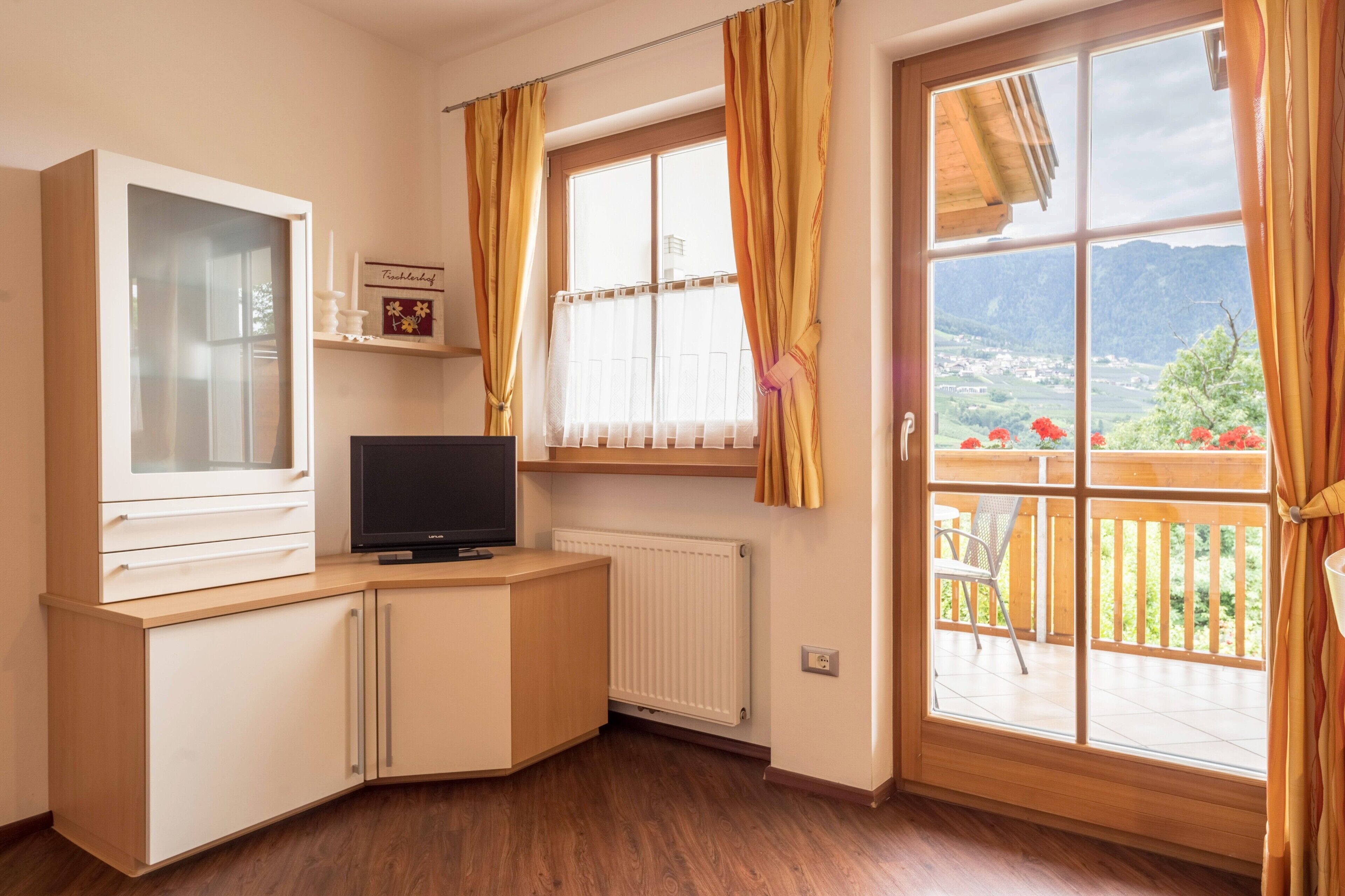 Ein Zimmer mit Fernseher auf einem Holzkasten, einem fenster mit Vorhängen, einer Heizung und Blick auf einen Balkon mit einem Stuhl.
