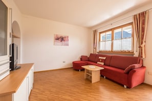 A modern living room with a red sofa, a small wooden table, a painting on the wall, and a window with curtains.