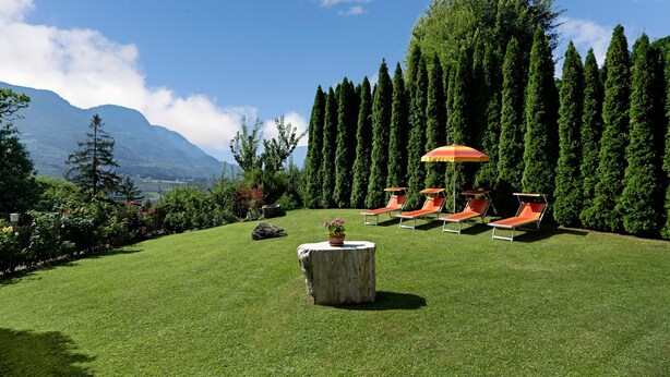 A well-maintained lawn with a round wooden table and a potted plant, surrounded by orange lounge chairs and a red umbrella.