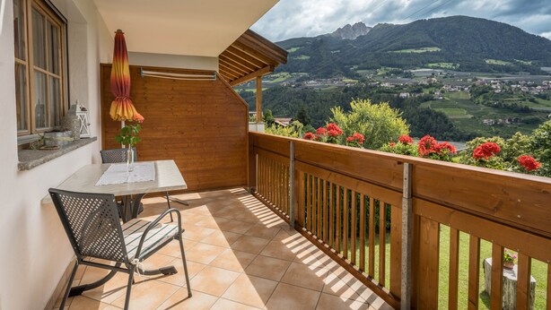 Un balcón con mesa y sillas, baranda de madera y vistas a un valle con montañas.