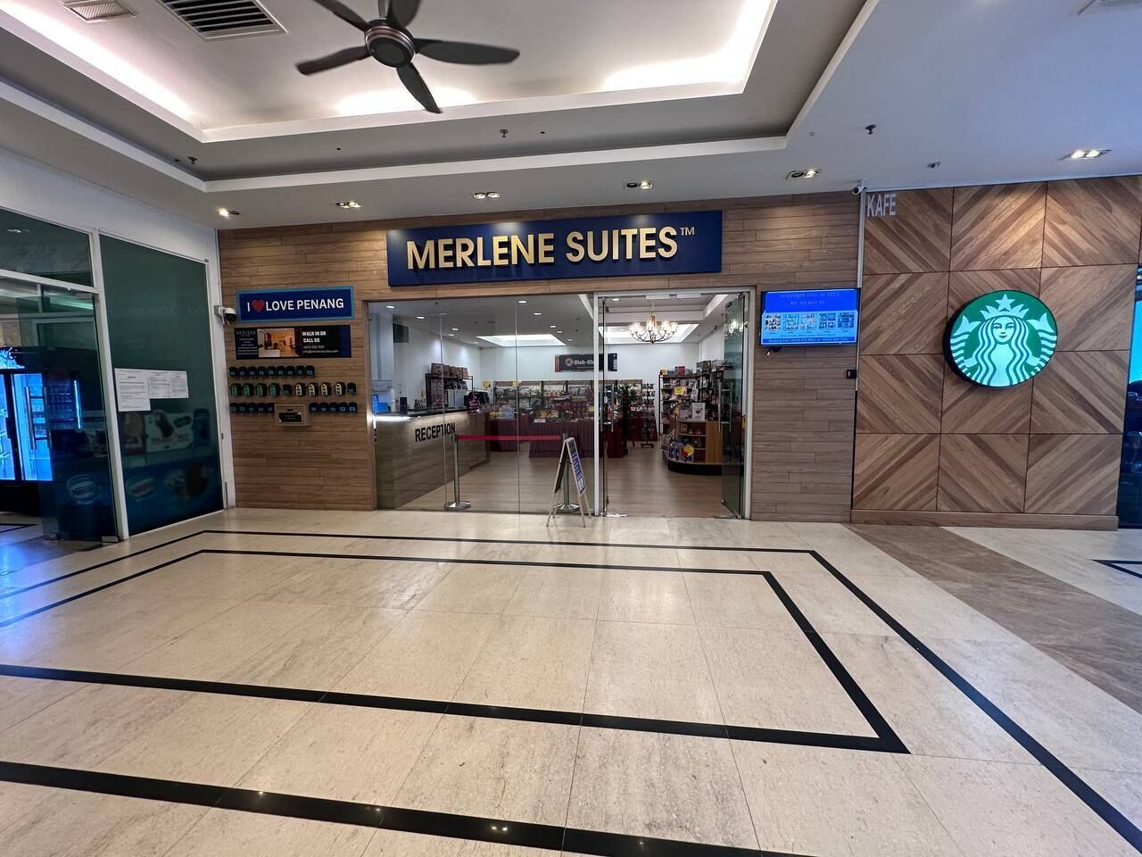 A hotel entrance with a reception desk, a sign reading "MERLENE SUITES™", and a Starbucks logo on the wall.