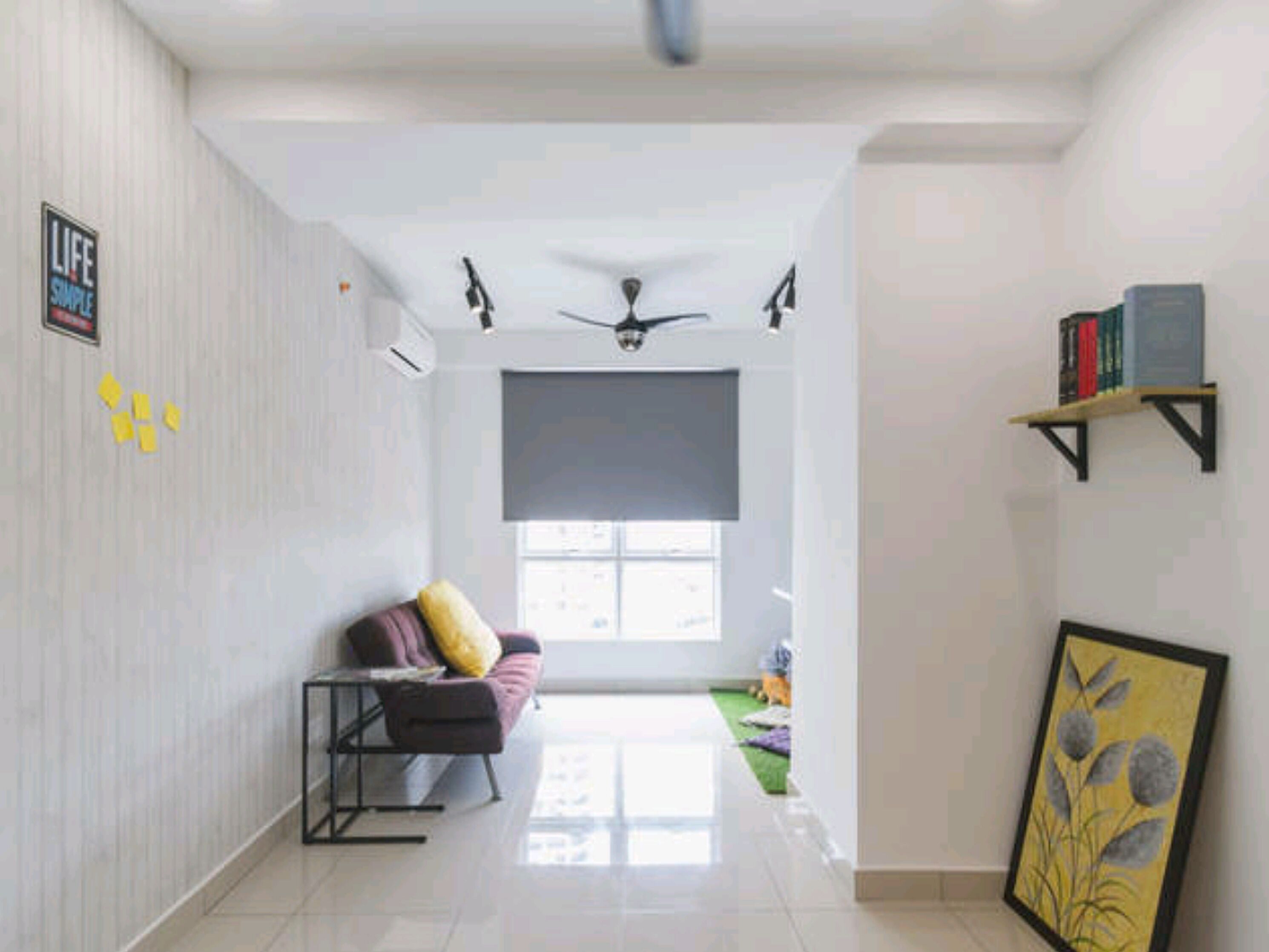 A spacious living area with a sofa, a small table, and a ceiling fan. There is a window with a view, a bookshelf, and a framed picture on the wall.