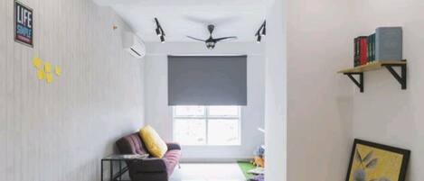 A spacious living area with a sofa, a small table, and a ceiling fan. There is a window with a view, a bookshelf, and a framed picture on the wall.