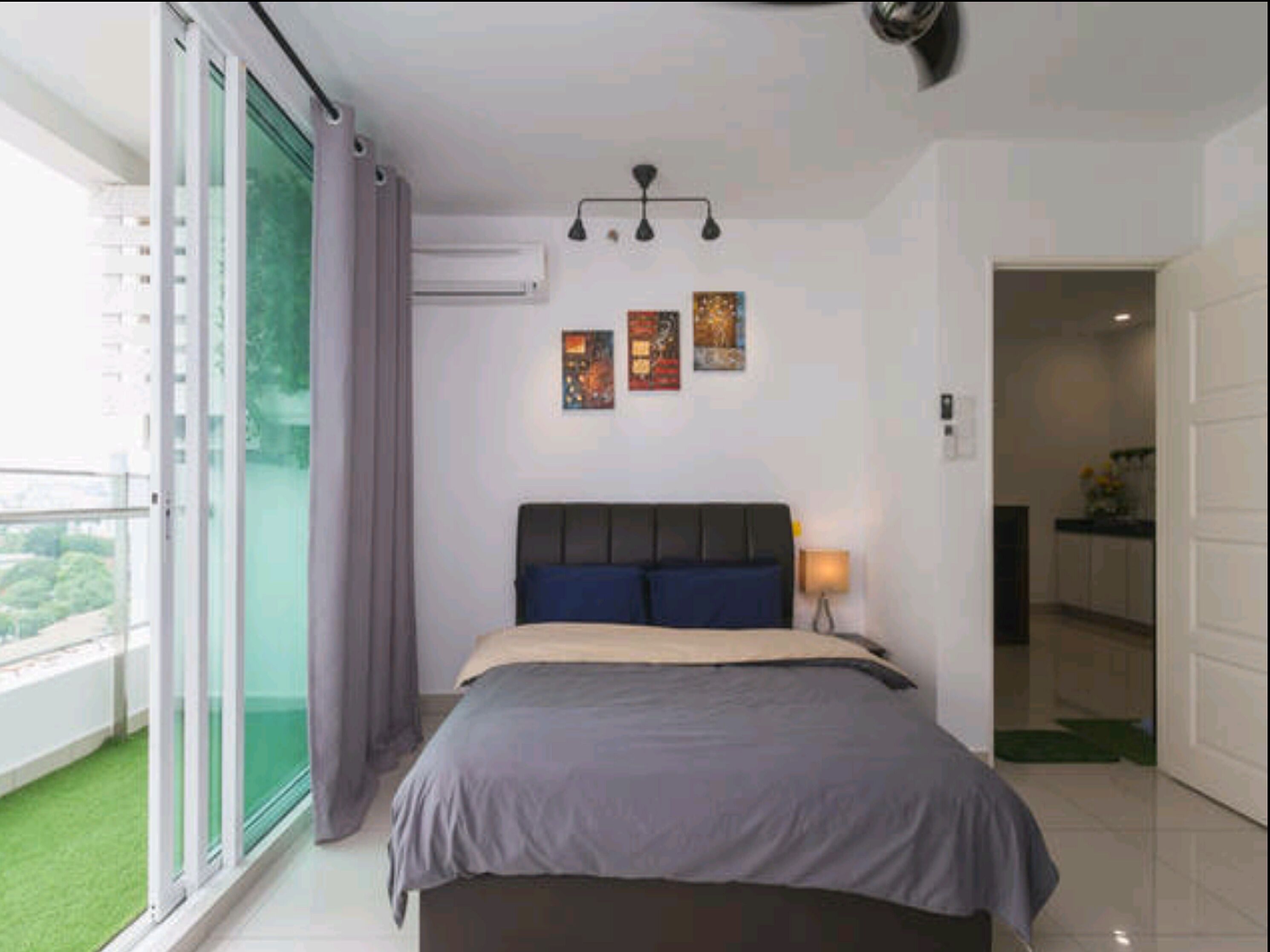 A modern bedroom with a large bed, a view of a balcony, and a kitchen visible through an open door.