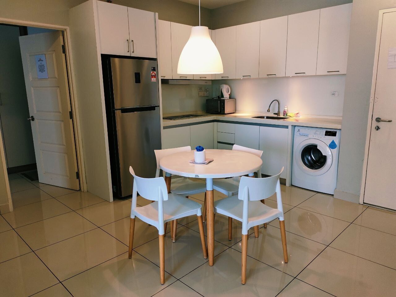 A compact kitchen with white cabinets, a stainless steel refrigerator, a washing machine, and a round table with chairs.