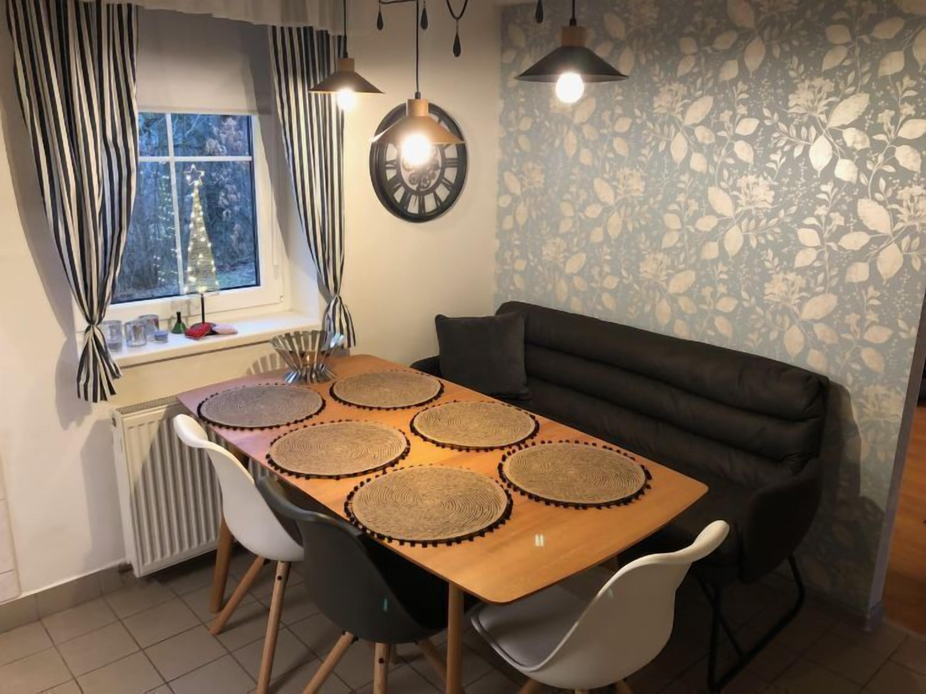 Un área de comedor con una mesa de madera, cuatro individuales tejidos, dos lámparas colgantes, un sofá negro y una ventana con cortinas.