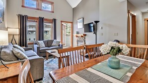 A modern living room with a wooden dining table, grey sofas, and a large window with curtains. - Close To It All! Rocky Mountain 2BR Loft Penthouse - Resort w Pool & 3 Hot Tubs! (Canmore)