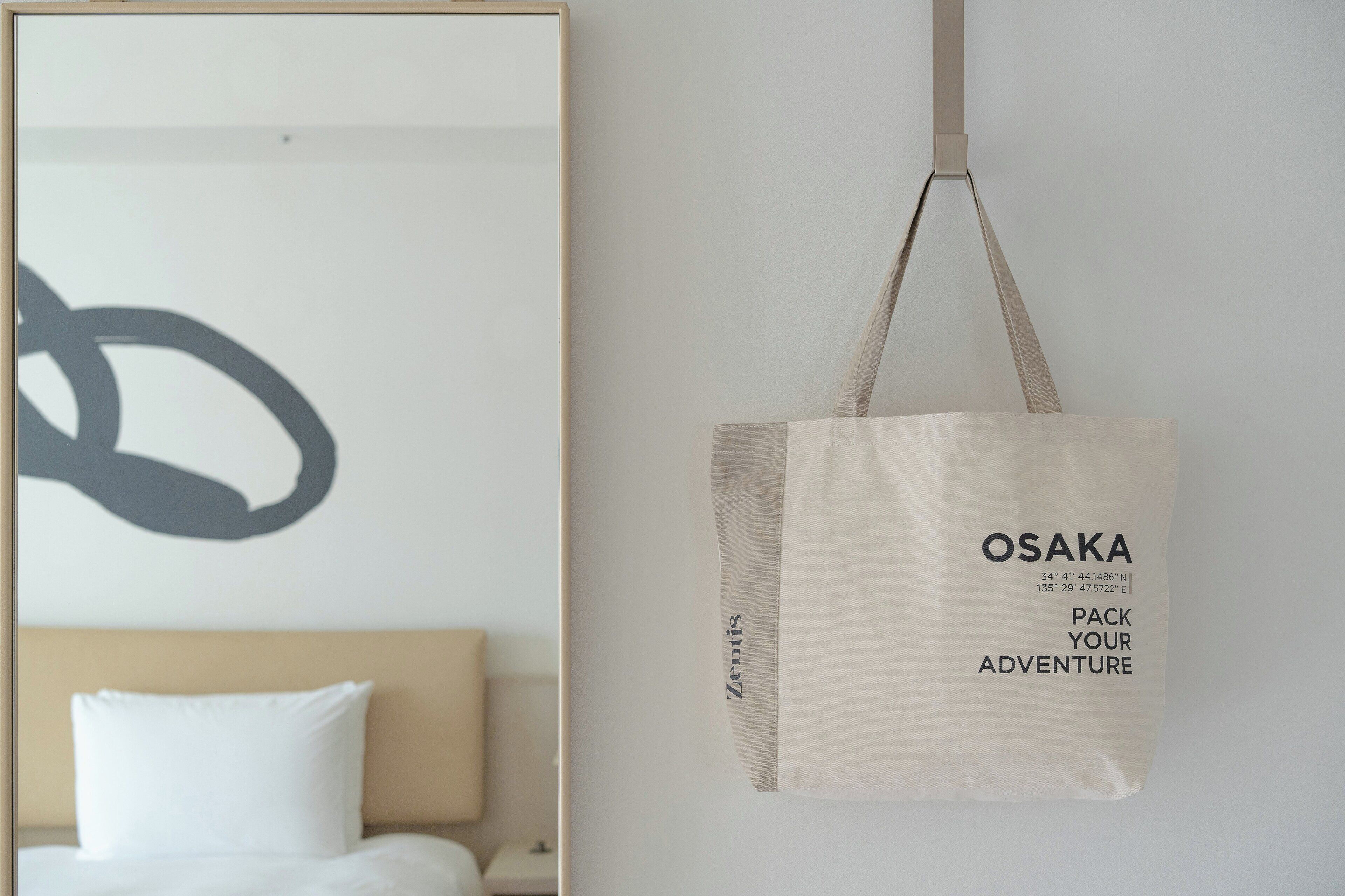 A hotel room with a bed, a wall with a large abstract artwork, and a hanging canvas bag with the text "OSAKA PACK YOUR ADVENTURE".