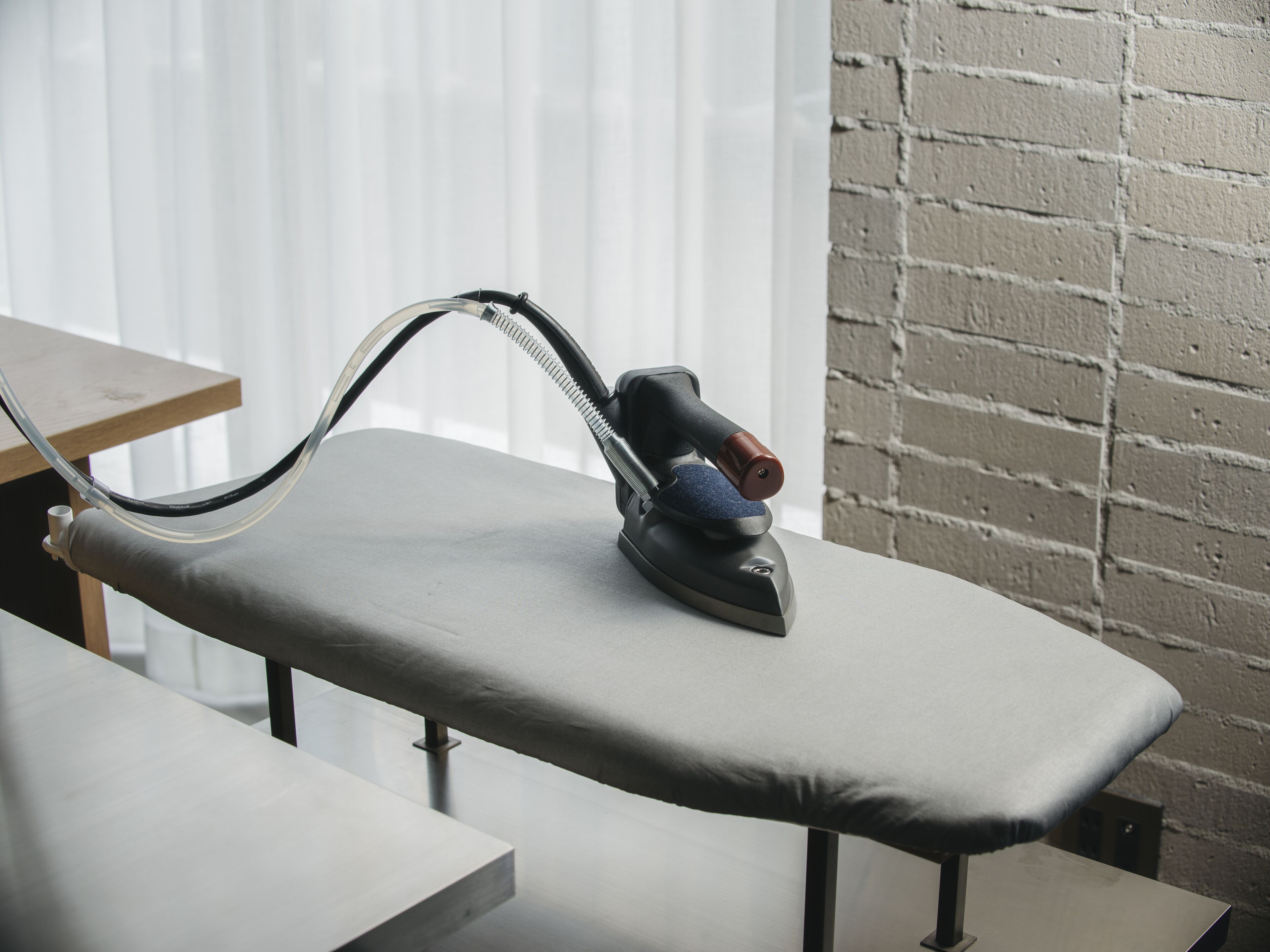 An ironing board with an iron on it, a wooden table, and a brick wall in the background.