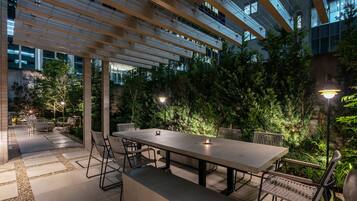 An outdoor patio with a long table and chairs, surrounded by lush greenery and a modern building in the background.
