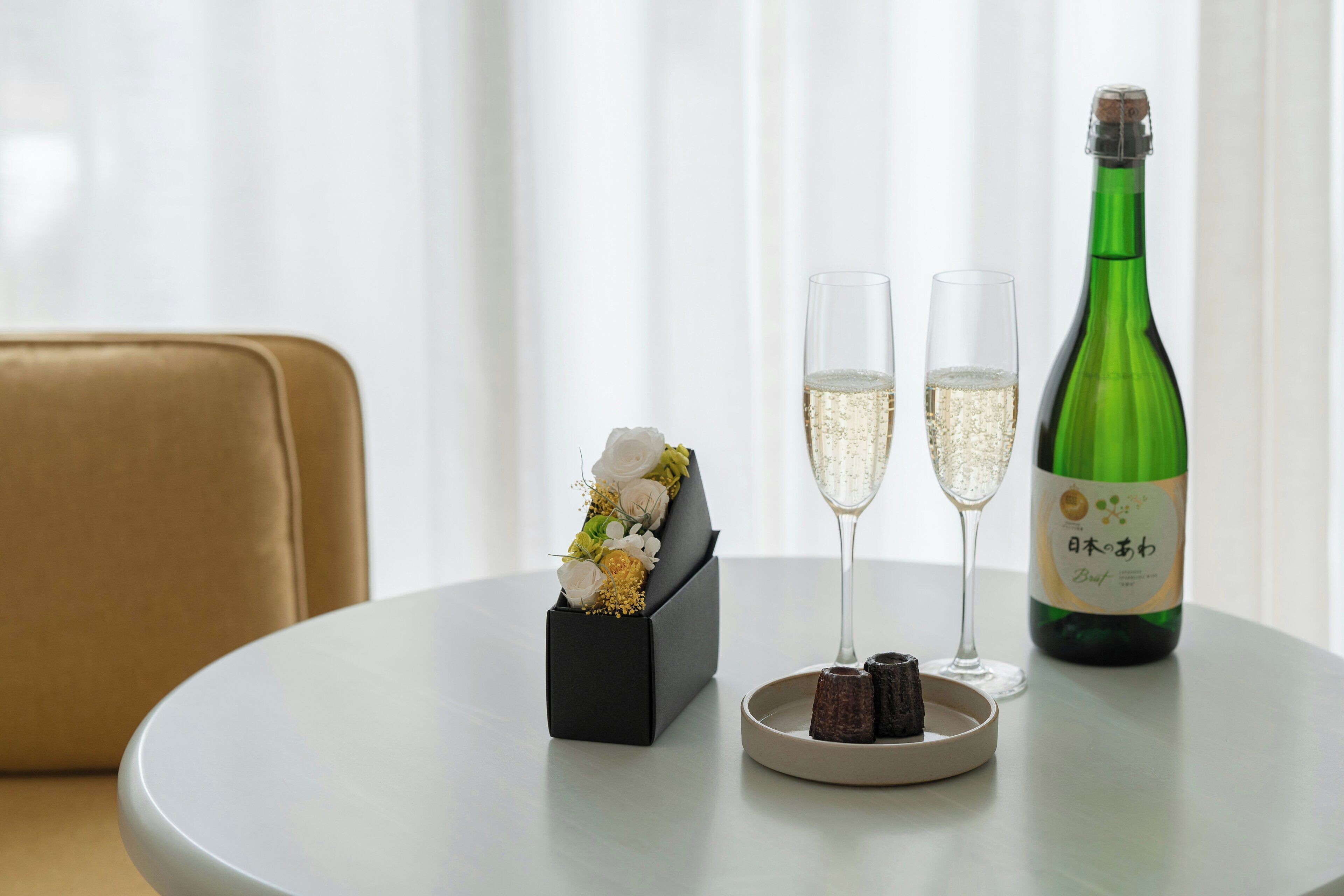 A round table with a bottle of champagne, two glasses, a box with flowers, and a plate with chocolates.