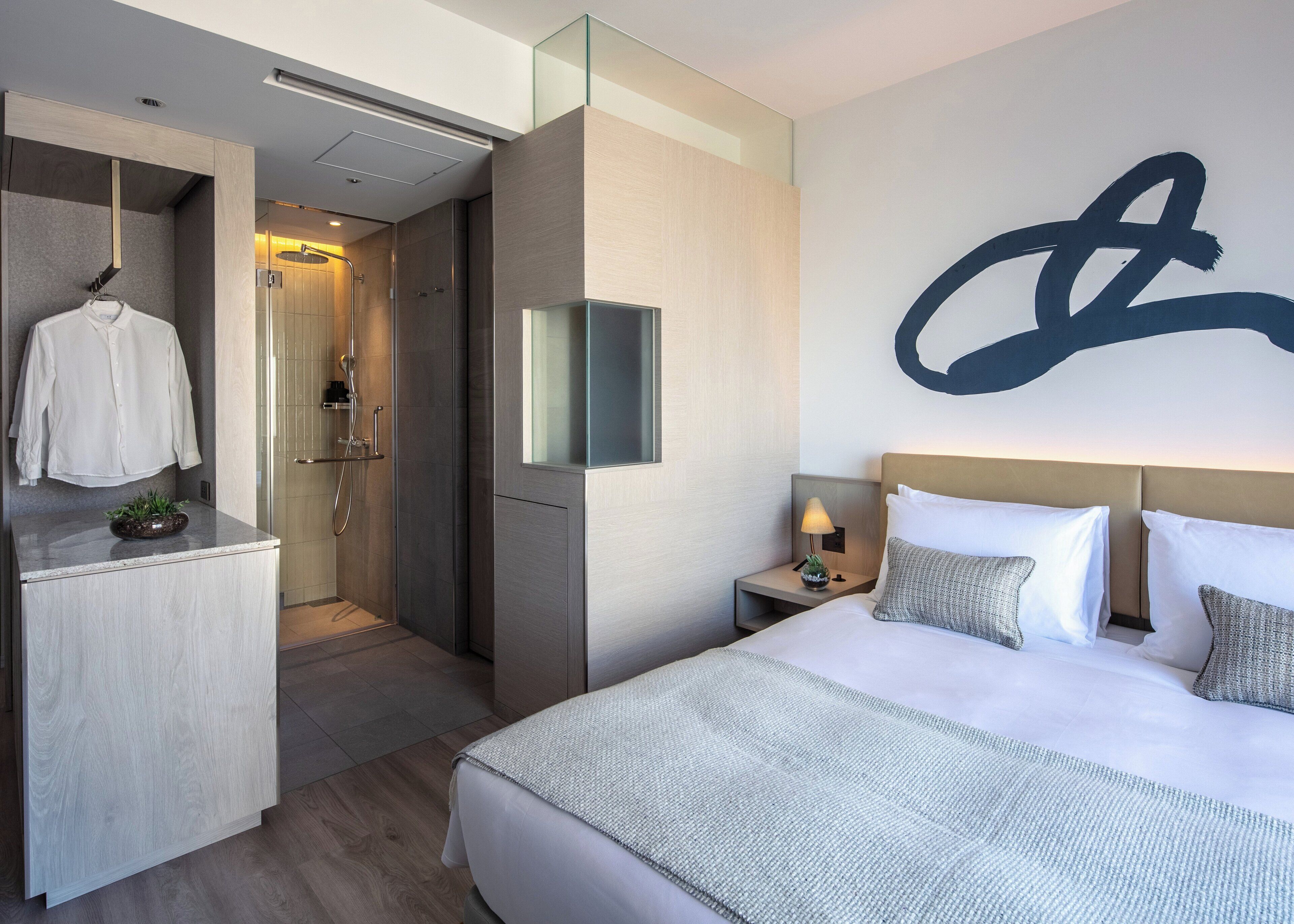 A modern hotel room with a large bed, a bedside table with a lamp, a mirror, and a shower area.