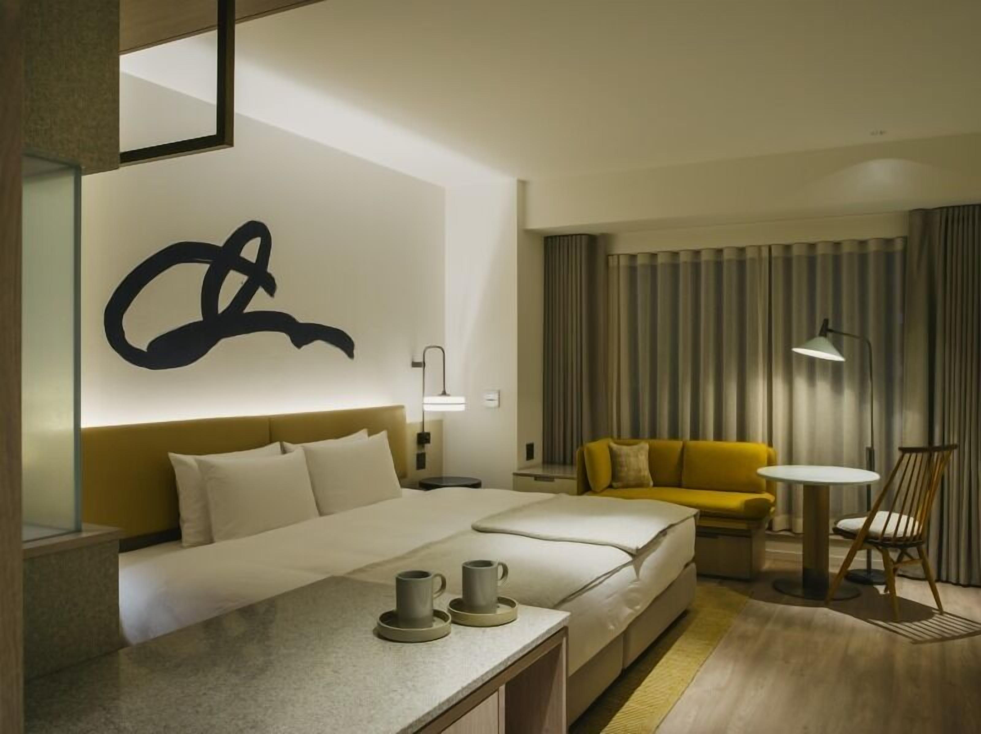 A hotel room with a bed, a sofa, a small table with cups, a lamp, and a large abstract wall art piece.