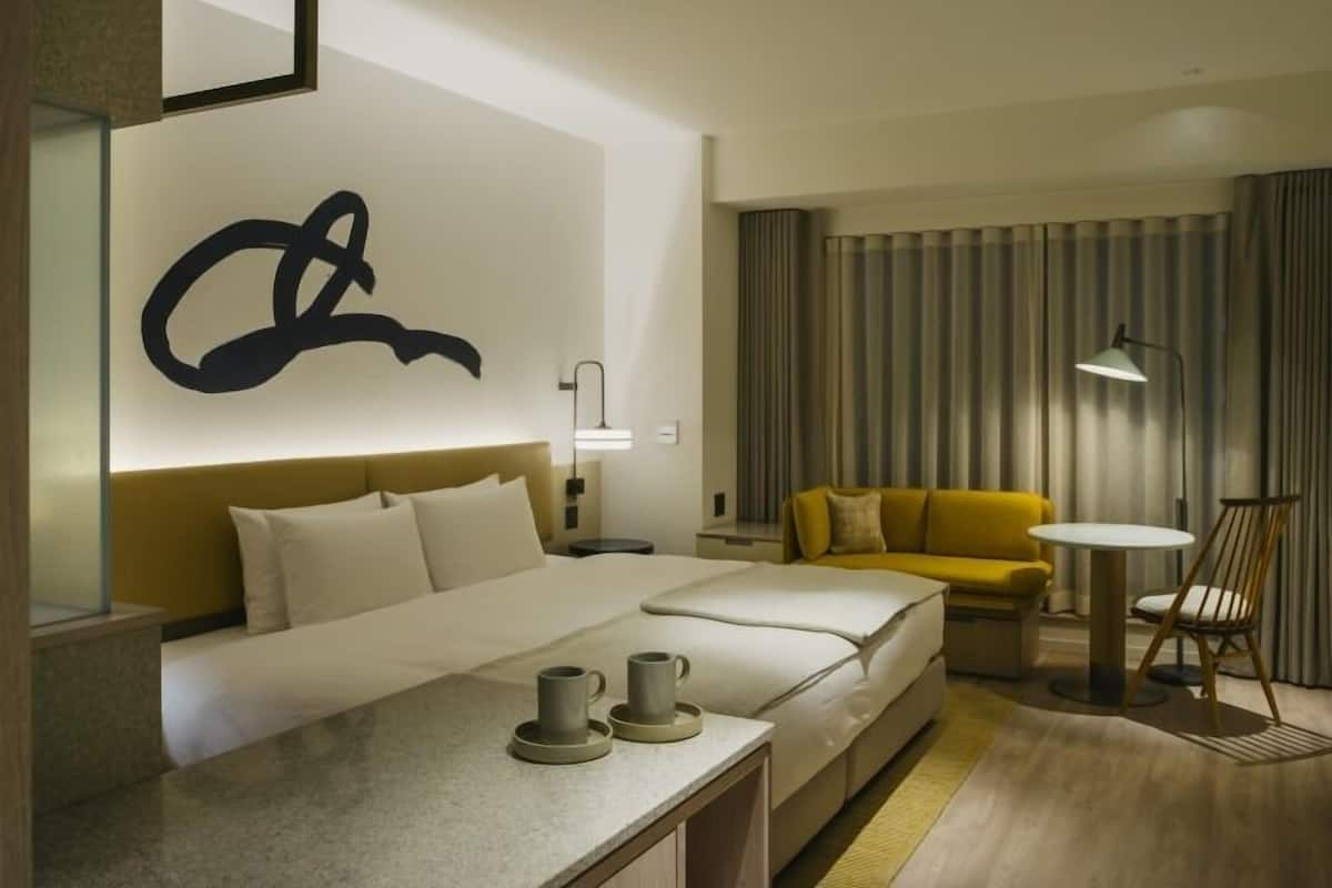 A hotel room with a bed, a sofa, a small table with cups, a lamp, and a large abstract wall art piece.
