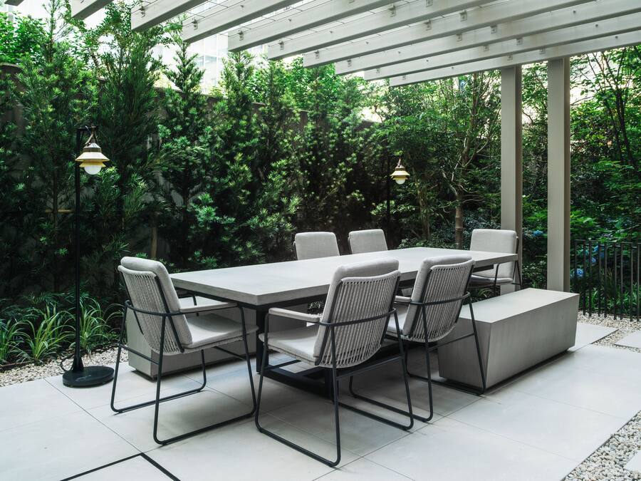 An outdoor patio with a rectangular table and six chairs, surrounded by lush greenery and a pergola structure.