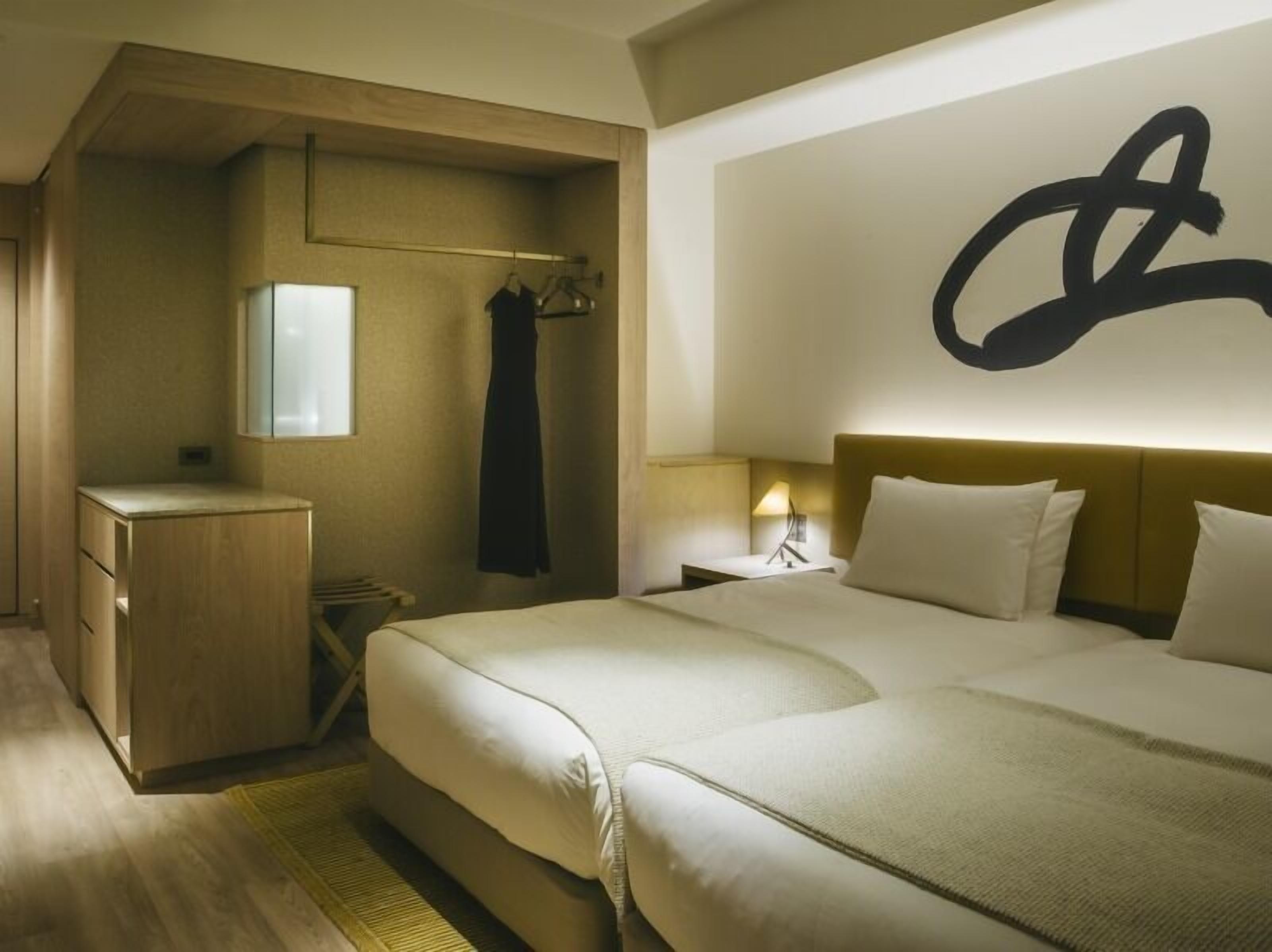 A hotel room with a bed, bedside table, lamp, wardrobe, and a large abstract wall art piece.