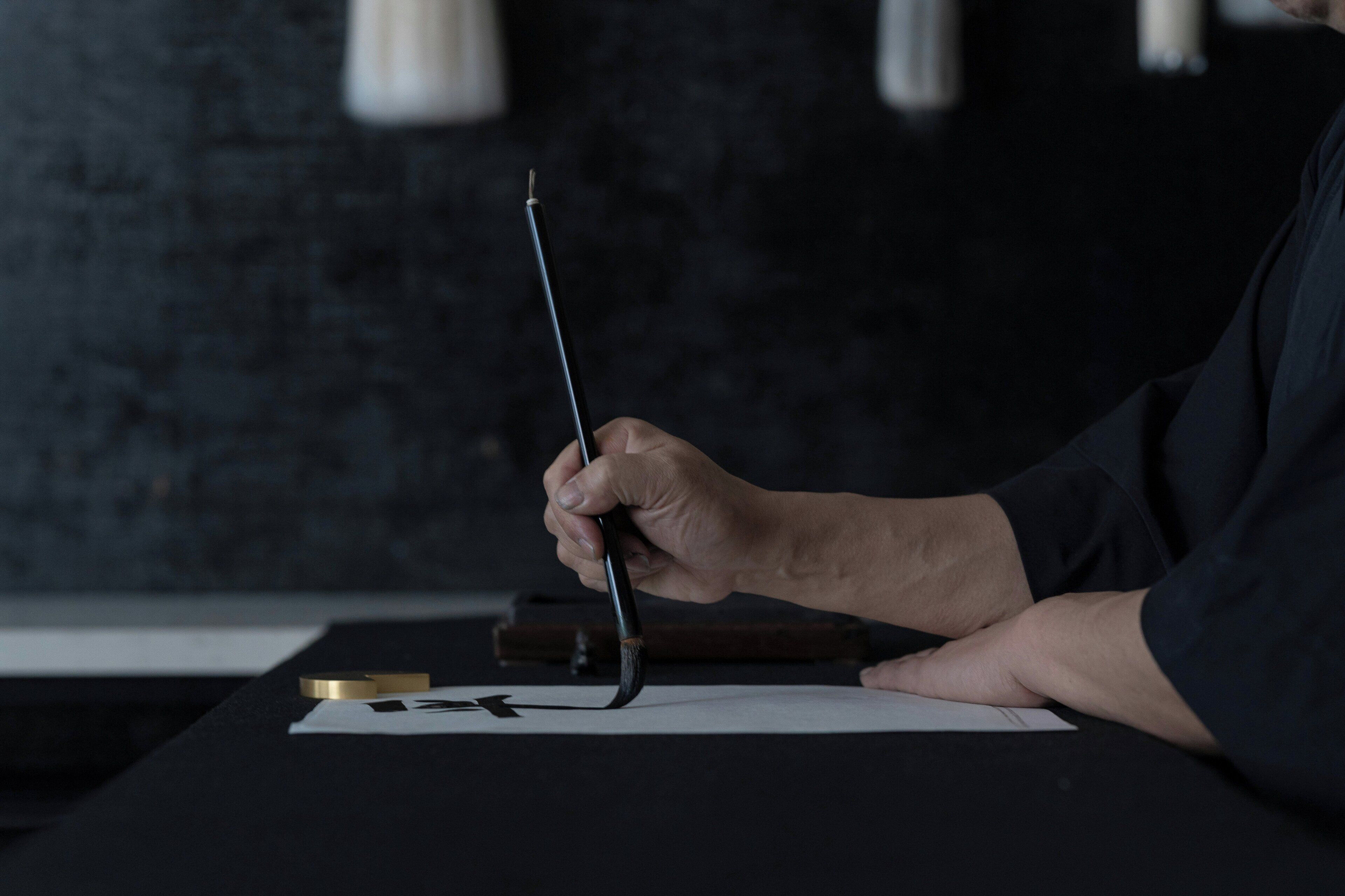 A person holding a brush, with a piece of paper and a gold object on a table.