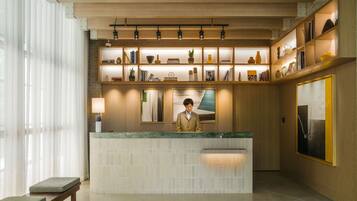 A reception desk with a wooden counter and a person standing behind it. There are shelves above the desk with various items, and a large painting is visible on the wall.