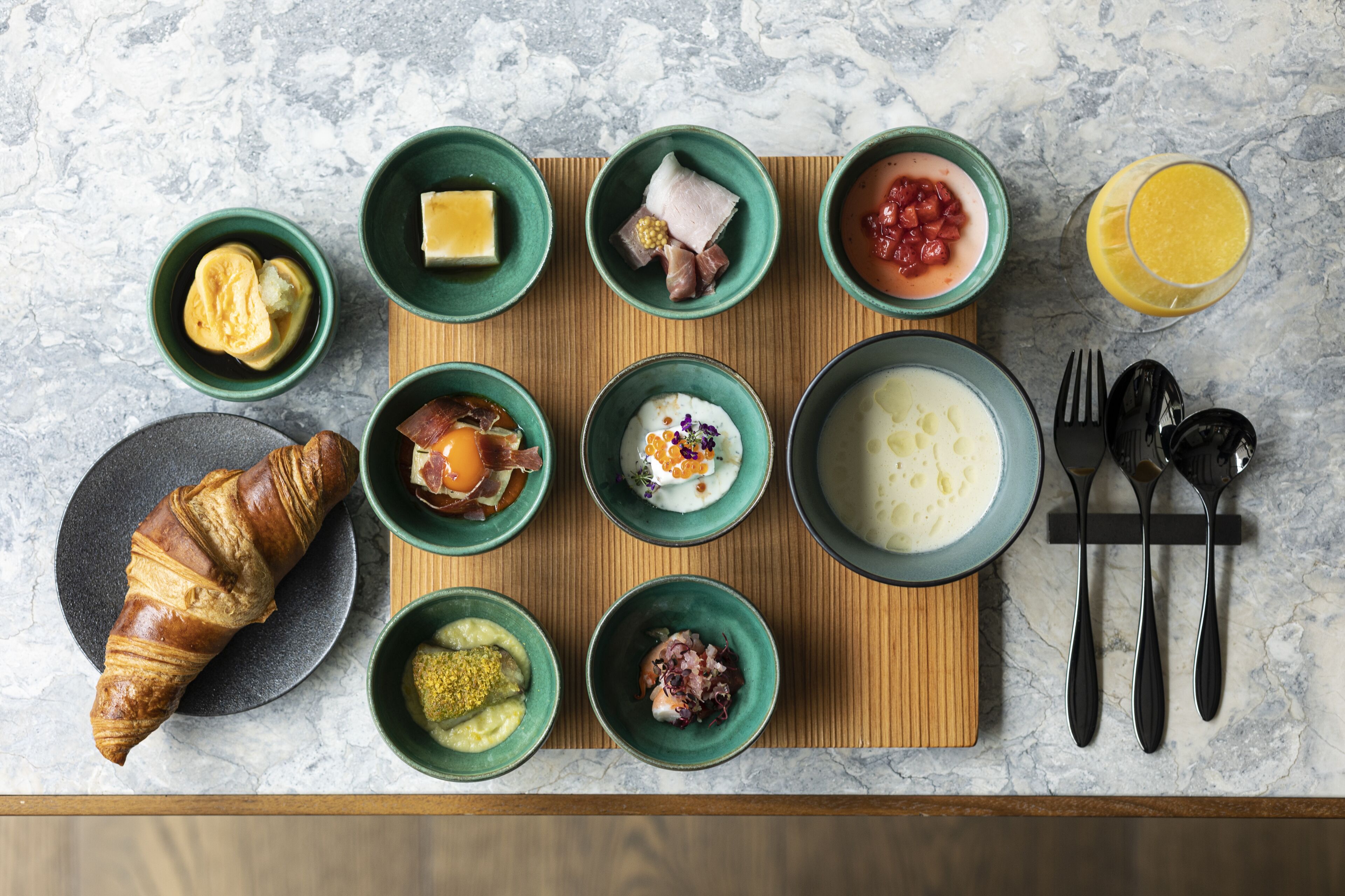 A breakfast spread on a wooden board with various dishes and a croissant.