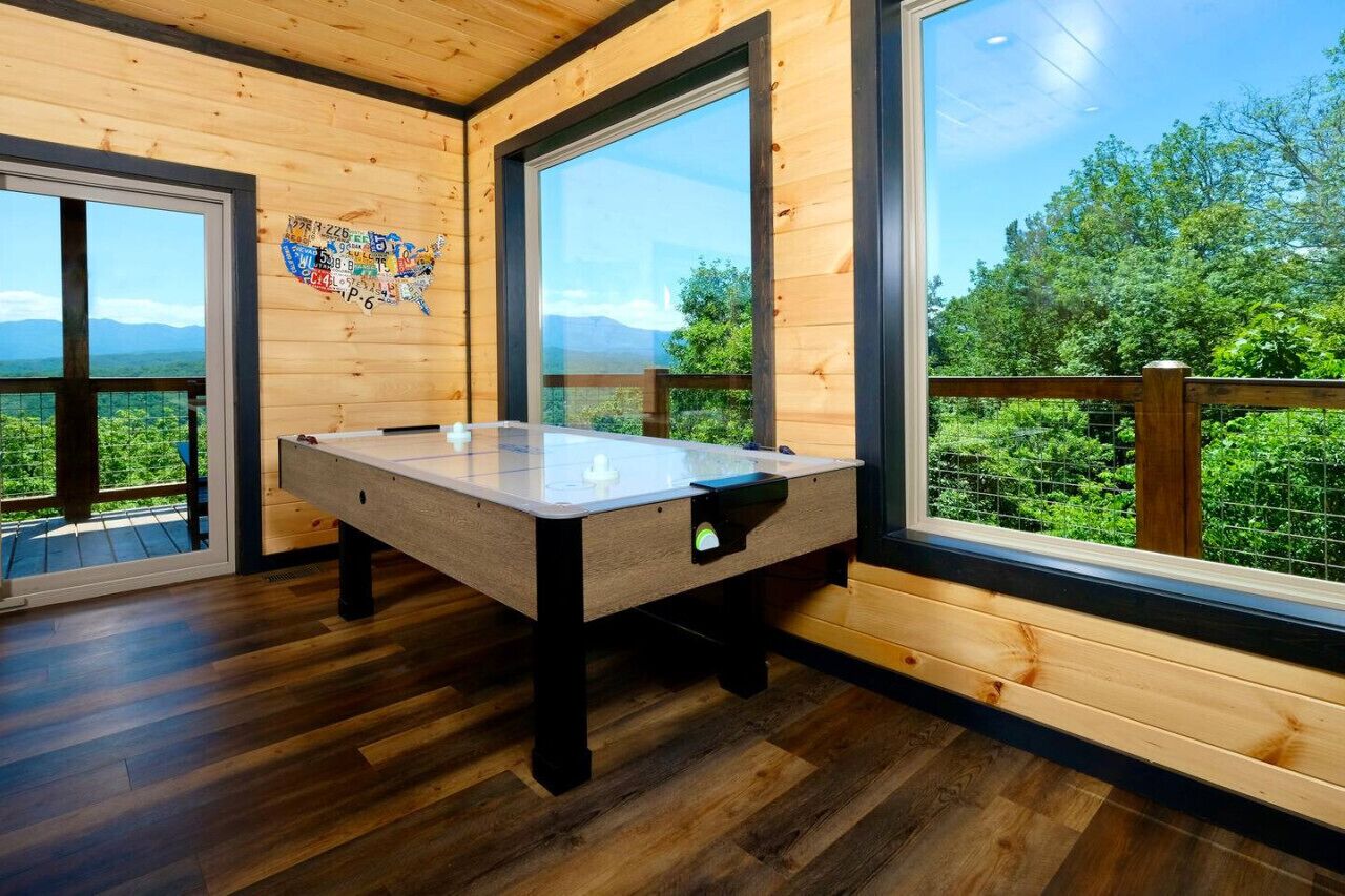 A wooden-floored room with a pool table, large windows, and a view of greenery.