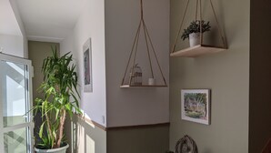Quarto com uma grande planta em vaso perto de uma janela, uma pequena mesa de madeira com um candeeiro e um armário fixado na parede com uma planta e uma gaiola de pássaros.