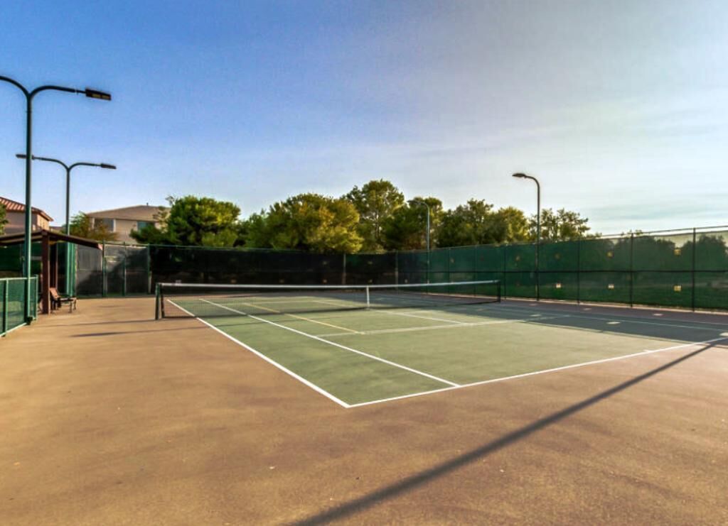Una cancha de tenis al aire libre con red, rodeada por una valla y farolas.