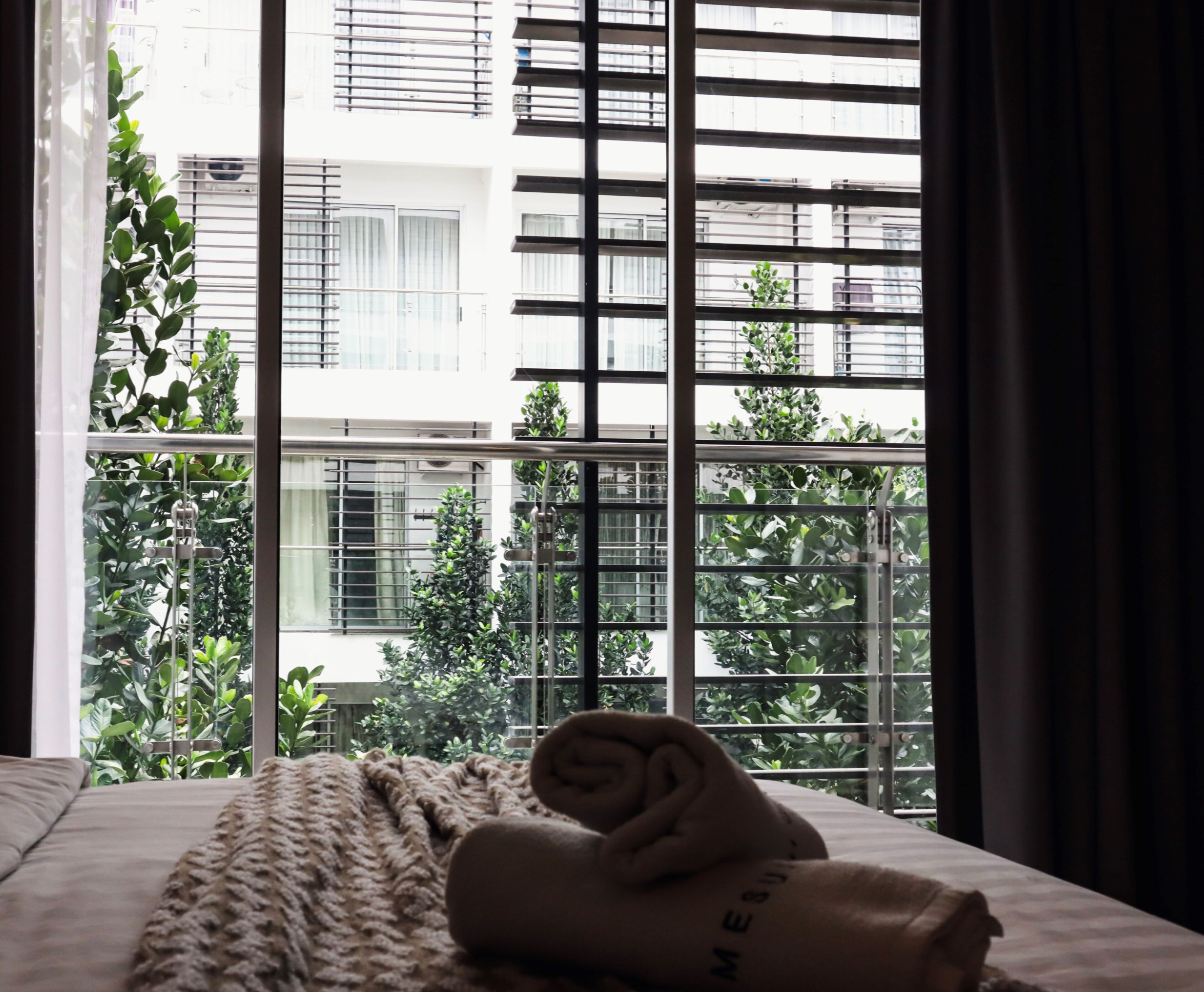 Habitación de hotel con cama, toalla enrollada y ventana que ofrece vistas a un edificio y follaje.
