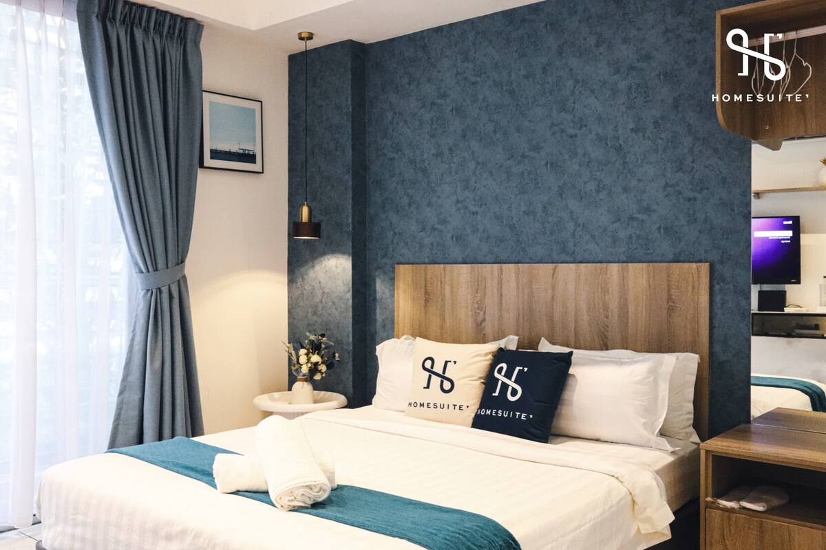 Une chambre d’hôtel avec un grand lit, une tête de lit en bois, une table de chevet, une télévision et une fenêtre avec des rideaux.
