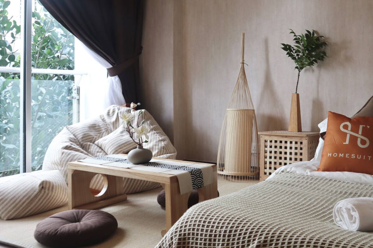 Une chambre bien éclairée, avec un lit, des oreillers, une table de chevet, une lampe et une fenêtre avec des rideaux.