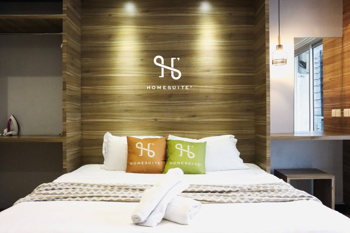 Une chambre d’hôtel avec une tête de lit en bois, deux oreillers portant la marque « HOMESUITE », un lit bien fait avec une couvre-lit à motifs, et une petite table de chevet avec une lampe.