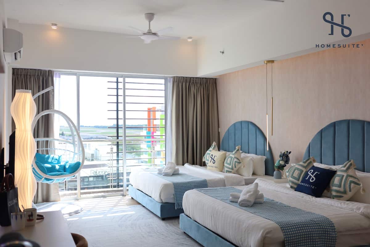 Une chambre d’hôtel avec deux lits, un fauteuil suspendu, une grande fenêtre avec des rideaux et un ventilateur de plafond.