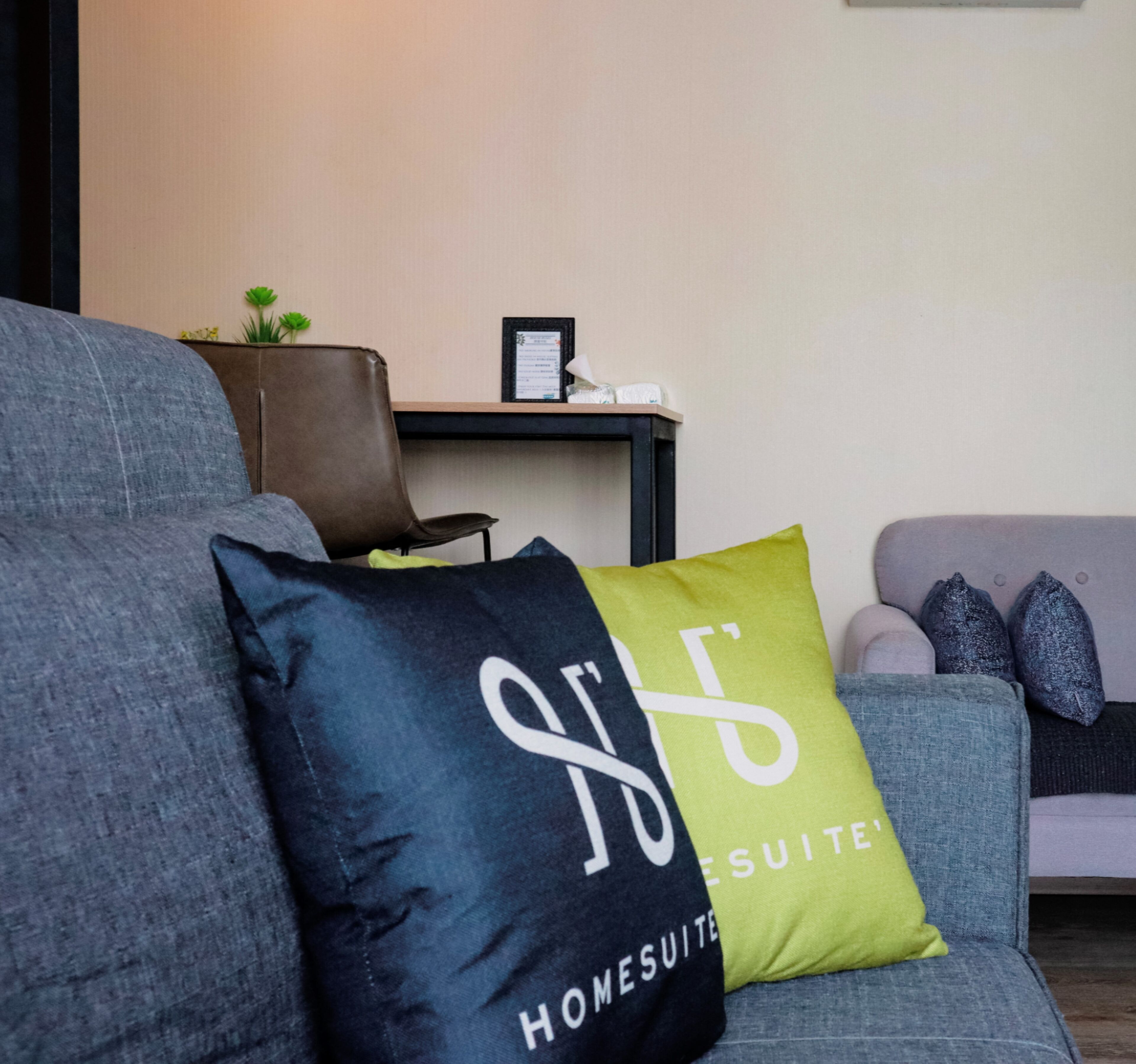 A modern living room with a grey sofa, a black and green pillow with the text "HONES SUITE," and a small table with a framed certificate.