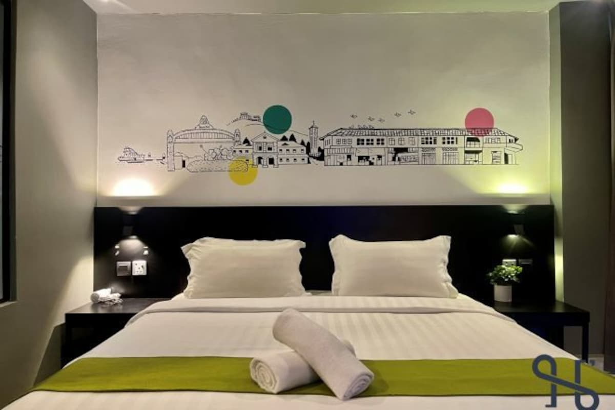 Quarto de hotel com cama, duas mesas de cabeceira, um mural na parede e uma mesa de apoio com abajur.