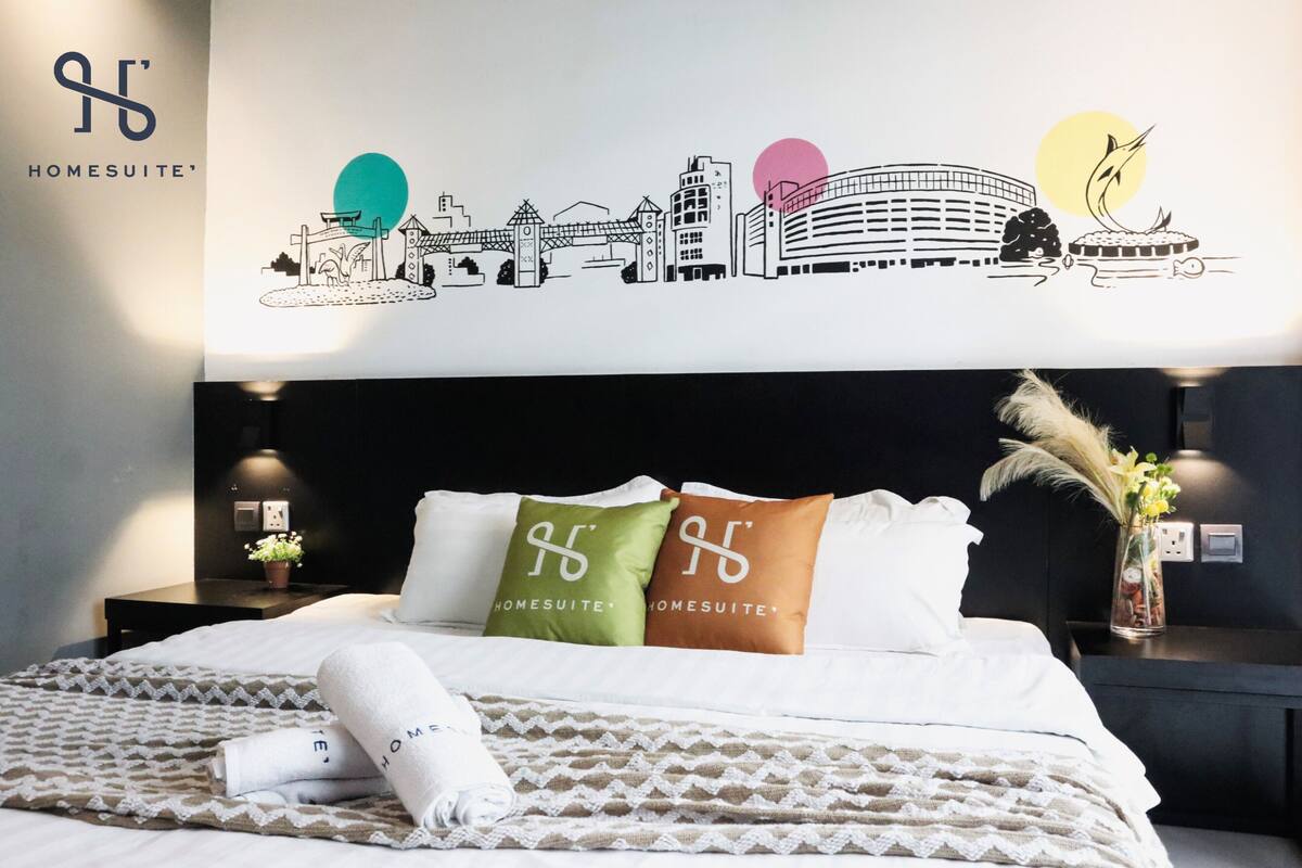Une chambre d’hôtel avec un lit, des oreillers, une table de chevet, un vase avec des fleurs et un motif décoratif sur le mur.