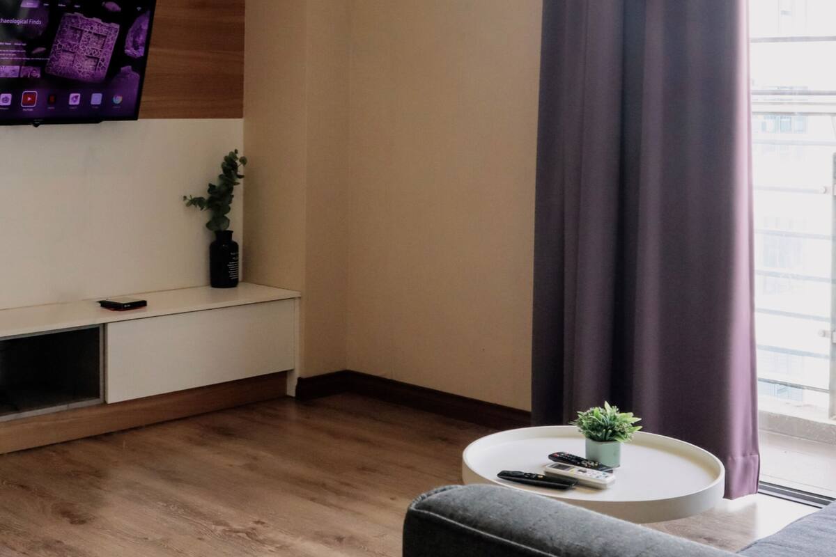 Un salon moderne avec un téléviseur à écran plat fixé au mur, un meuble de rangement blanc, une table basse ronde avec une télécommande, un canapé gris et une fenêtre avec des rideaux.