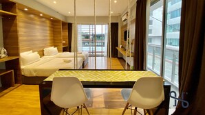 A hotel room with two beds, a dining table with chairs, a TV, and a balcony with a view of buildings.