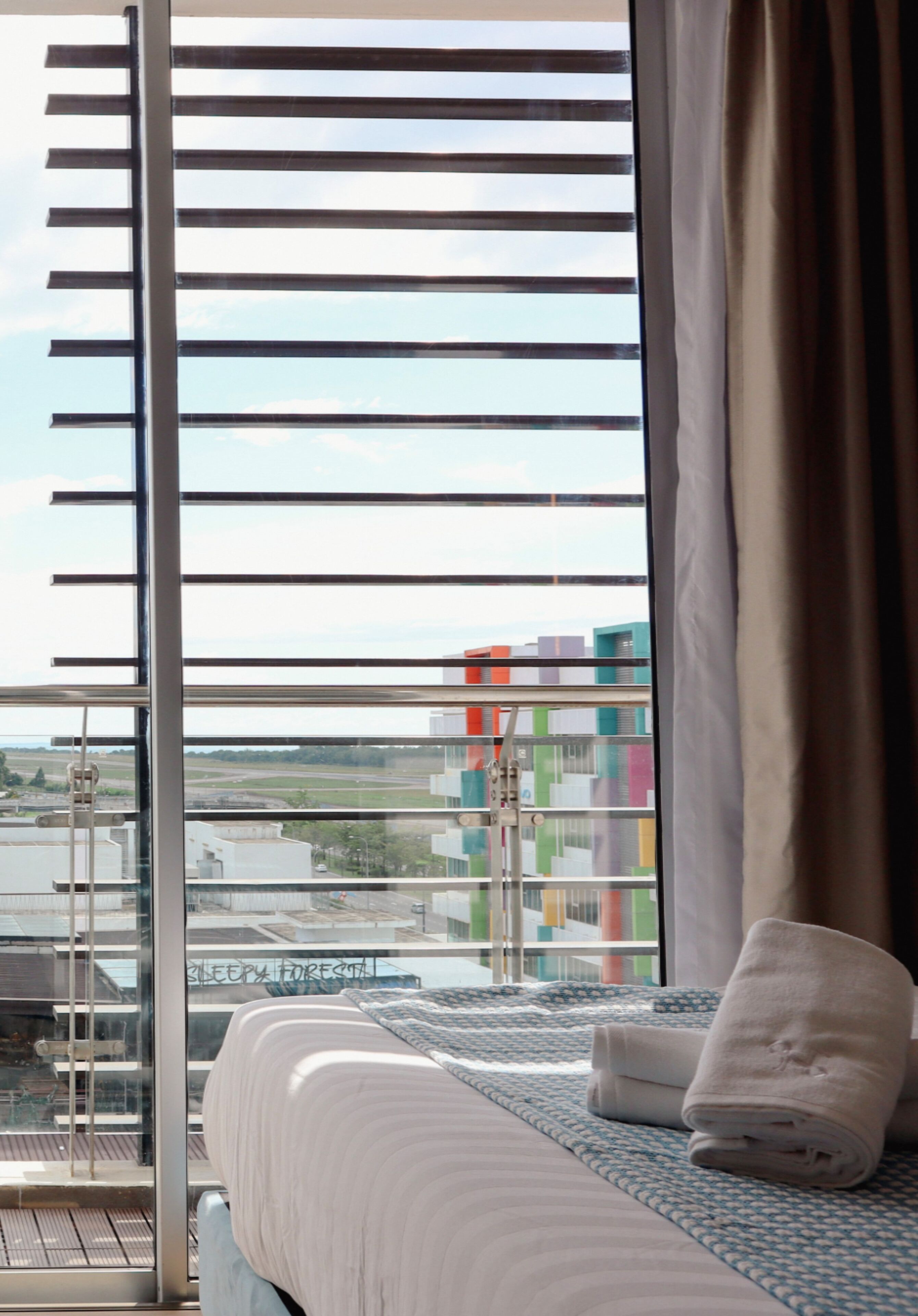 A hotel room with a bed, a window with horizontal blinds, and a view of colorful buildings outside.