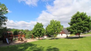 A playground with swings and a slide, surrounded by green grass and trees.